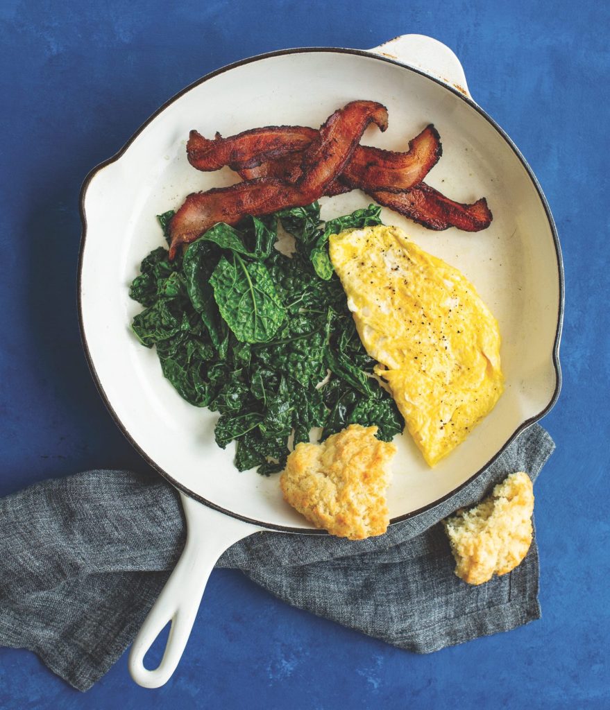 scrambled eggs, bacon, greens, and a biscuit in a cast-iron skillet