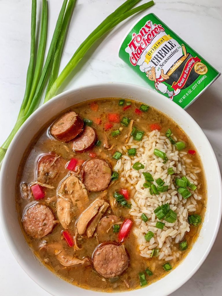 Bowl of Trinity Gumbo, flanked by stalks of green onions and Tony Chachere's Original Seasoning