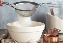 Flour Power Flour sifting into a bowl. A whisky, stack of measuring cups, and glass container of flour huddled close.