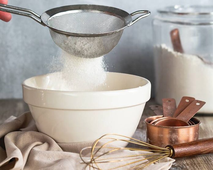 Flour sifting into a bowl. A whisky, stack of measuring cups, and glass container of flour huddled close.