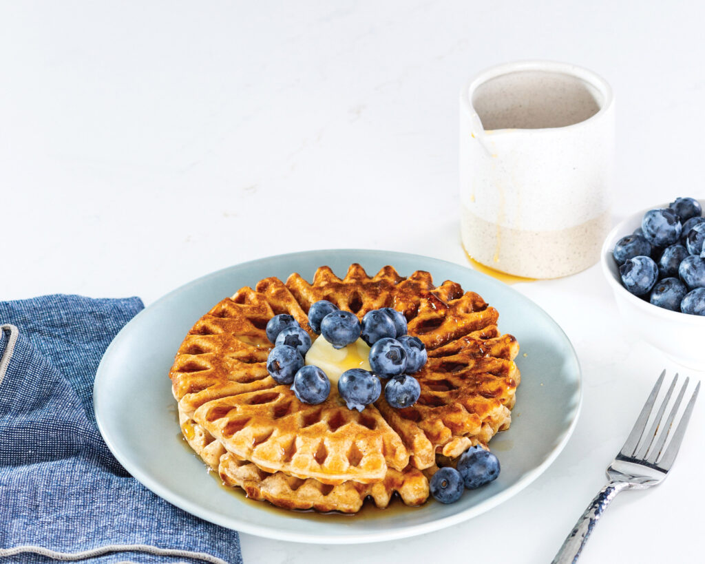 Blueberry waffles with syrup on a light blue plate beside a bowl of blueberries.